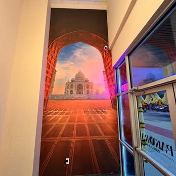 Pakora Indian Eatery - A mural depicting the Taj Mahal is painted on a tall interior wall, featuring a detailed image of the monument against a sky with pink and blue hues. To the right, there is a glass door with a metal frame, decorated with yellow flowers, and a security camera is mounted near the top of the mural. The floor is black with a marble pattern, and the ceiling has a grid of white tiles. - Indian Near Me - Irving, Texas