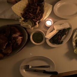 Full Chicken // Fries // Broccolini with Feta // Caesar Salad - A dinner table with a variety of Mexican dishes, including a bowl of grilled chicken, a basket of French fries, a small bowl of green salsa, grilled vegetables with cheese, and a salad. The table is dimly lit with a small candle, and there are plates, utensils, and a napkin visible.