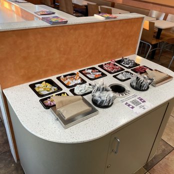 Taco Bell - A restaurant condiment station features a curved, speckled countertop with a brown partition behind it. The station holds several small black trays filled with condiment packets, including ketchup, mustard, and other sauces. There are stacks of napkins and a trash bin integrated into the station. The background shows dining chairs and tables, indicating a casual dining environment. - Fast Food Near Me - Middleton, Wisconsin
