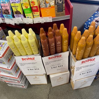 BevMo! - The image shows two cardboard displays containing bottles of whipped vodka, one vanilla flavored and the other mocha flavored, each with a 200 ml capacity. The bottles are arranged upright, with the vanilla flavor on the left and mocha in the middle. Promotional signs indicate a deal of three bottles for $33. Behind the displays, there are shelves with various snack and beverage products, including energy drinks and bars, in a retail store setting. The floor is concrete with visible cracks. - Beer, Wine & Spirits Near Me - Temecula, California