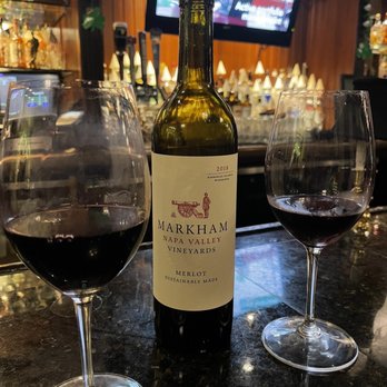 McCarthy's Pub - Excellent wine menu - A 2018 Markham Napa Valley Merlot wine bottle is centered on a dark bar counter, with two glasses of red wine on either side. The background features a bar with taps and a television screen showing a person in a suit. The setting suggests a bar or restaurant environment with a focus on wine service. - Irish Pub Near Me - Tequesta, Florida