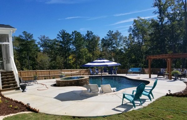 A backyard with a swimming pool featuring a circular spa, shaded seating area with an umbrella, surrounding lounge chairs, a wooden fence, and lush trees in the background.