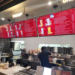 A barista is working behind a counter in a boba tea shop, with digital menu screens displaying various milk teas and fruit teas.