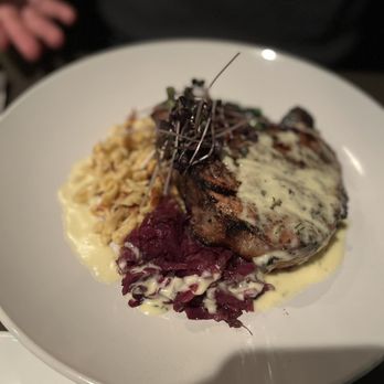 C3 Bar - Pork chop - A white plate holds a serving of grilled meat topped with a creamy herb sauce, accompanied by mashed potatoes and a portion of cooked red cabbage. The dish is presented on a dark table at C3 Bar in Bloomington, IN, which offers a diverse menu and seasonal cocktails. - Cocktail Bars Near Me - Bloomington, Indiana