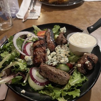 Teddy G's - A salad featuring grilled meat, blue cheese crumbles, sliced cucumbers, tomatoes, and red onions, served with a side of creamy dressing on a black plate. - Pubs Near Me - Sturbridge, Massachusetts