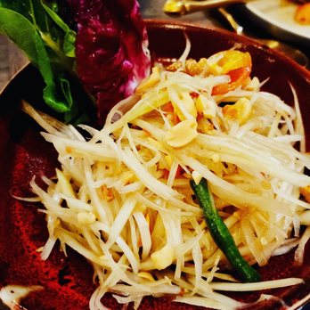 The Nuaa Table - Green papaya salad - A bowl containing green papaya salad with shredded papaya, peanuts, and vegetables, served with leafy greens on the side. - Thai Near Me - Brooklyn, New York