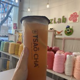Grapefruit and Peach Jasmine Tea - A person holds a large cup of tea labeled 'TSÃO-CHA' inside a tea shop decorated with yarn spools and plush wall art.