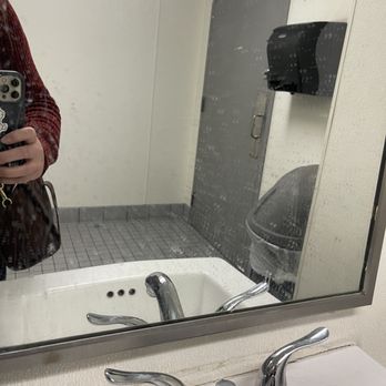 Staples - A bathroom mirror with water spots reflects a white sink with a chrome faucet, a person holding a phone in the left side of the frame, and a bathroom door with a handle and a black paper towel dispenser. The mirror shows a trash can with a plastic liner and a tiled floor in the background. - Shipping Centers Near Me - Downey, California