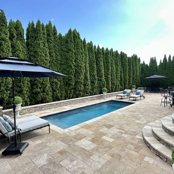Gappsi, INC. - The image shows a spacious backyard with a rectangular swimming pool at the center, bordered by a stone patio. There are lounge chairs with cushions and umbrellas for shade, along with outdoor dining furniture. Tall, dense green hedges provide privacy, and potted plants are placed near the house. The house has white siding and exterior lighting fixtures. The sky is clear with some clouds, indicating a sunny day. - Masonry/Concrete Near Me - smithtown, New York