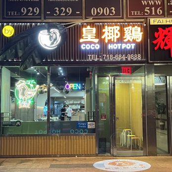Coco Hotpot - Storefront - The image shows the storefront of Coco Hotpot, a restaurant specializing in coconut-based hotpot dishes. The facade includes illuminated signs with Chinese characters and the English name, along with a phone number. A neon chicken logo is displayed on the glass window, which also reveals the interior with staff and seating. The entrance has a glass door with a small seating area visible inside, and a neon 'OPEN' sign is lit. - Hot Pot Near Me - Flushing, New York
