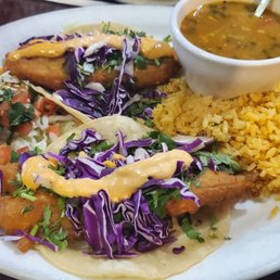 Fish Tacos - A plate of fish tacos with purple cabbage slaw and sauce, accompanied by yellow rice, a side salad with diced tomatoes and onions, and a cup of soup, served at Los Generales in Spring, TX. The meal is presented on a white oval plate with additional small bowls of sauce or soup in the background.