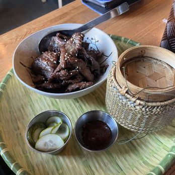 Thip Khao - A bowl of cooked meat with sesame seeds, served on a bamboo tray with pickles and a dipping sauce, in a restaurant setting. - Laotian Near Me - Washington, DC, District of Columbia