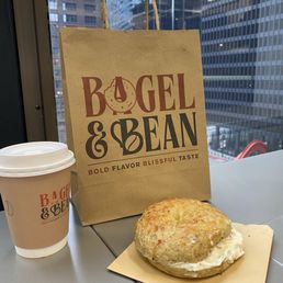 Rosemary and veg cream cheese - A bagel sandwich with cream cheese is set on a small paper plate on a table, with a coffee cup bearing the Bagel & Bean logo nearby. Behind the items, a paper shopping bag from Bagel & Bean features the logo and slogan. The background shows a cityscape with tall glass buildings, indicating an urban setting, likely inside a cafe or near a window.