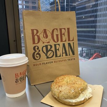 Bagel & Bean - Rosemary and veg cream cheese - A bagel sandwich with cream cheese is set on a small paper plate on a table, with a coffee cup bearing the Bagel & Bean logo nearby. Behind the items, a paper shopping bag from Bagel & Bean features the logo and slogan. The background shows a cityscape with tall glass buildings, indicating an urban setting, likely inside a cafe or near a window. - Coffee & Tea Near Me - Chicago, Illinois