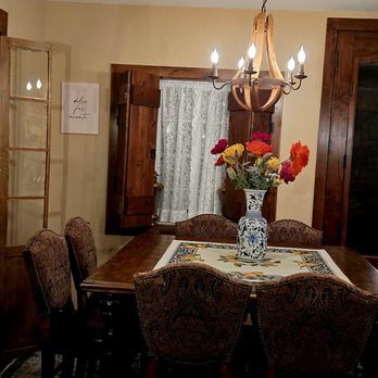 Level B - The image shows a dining room with a wooden table surrounded by six upholstered chairs with patterned fabric. A chandelier hangs above the table, and a large vase with colorful flowers is placed on a table runner. Wooden window shutters and a lace curtain are visible on the windows, and the room has warm lighting and decorative elements, including a stone wall on the left side. - General Contractors Near Me - Denver, Colorado