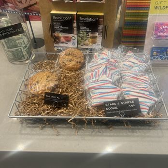 Wildflower - Special Stars & Stripes Cookie - A display on a counter features three cookies, two labeled as 'The Royal' and one in the middle, all wrapped in clear plastic. To the right, there are individually wrapped 'Stars & Stripes' cookies with red, white, and blue icing, priced at $3.99. Behind the display, there are tea bags and promotional boxes for tea or herbal infusions, along with various signs and packaging. The setting appears to be a cafe or retail store offering baked goods and beverages. - Sandwiches Near Me - Tempe, Arizona