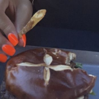 Empire - Burger - The image shows a person with orange-painted nails holding a French fry close to a large burger. The burger has a beef patty, lettuce, and is topped with a generous layer of chocolate sauce. It is served on a metal tray with some lettuce at the bottom. The person is wearing a dark top, and the focus is on the burger and hand. - Burgers Near Me - Naperville, Illinois