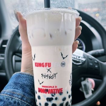 Kung Fu Tea - Coconut Milk Tea - A person wearing a denim jacket holds a large plastic cup of bubble tea with black tapioca pearls at the bottom. The cup has branding from Kung Fu Tea and Spirit, with the words 'Principle 2: Innovative' printed on it. The background shows the interior of a vehicle, including a steering wheel and a rainy window outside. - Bubble Tea Near Me - Pasadena, Maryland