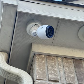 Data Source Solutions - Front camera - A white security camera is mounted on the underside of a building's eaves, facing outward. The camera is attached to a white circular mount. The building exterior includes a brick wall and a white drainpipe. The sky is visible in the background, indicating an outdoor setting. - Security Systems Near Me - Converse, Texas