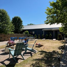 two people sitting at a picnic table - The image shows two people sitting on light blue outdoor chairs in a yard with a brick-paved area. They are under the shade of a tree, with a building in the background and a clear blue sky overhead. The yard has some small shrubs, a wooden fence, and outdoor lighting along a pathway. The scene appears to be a relaxed outdoor setting, possibly at a business or community space.