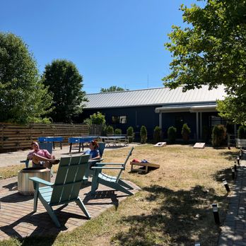 SideYard - two people sitting at a picnic table - The image shows two people sitting on light blue outdoor chairs in a yard with a brick-paved area. They are under the shade of a tree, with a building in the background and a clear blue sky overhead. The yard has some small shrubs, a wooden fence, and outdoor lighting along a pathway. The scene appears to be a relaxed outdoor setting, possibly at a business or community space. - Bars Near Me - Sawyer, Michigan