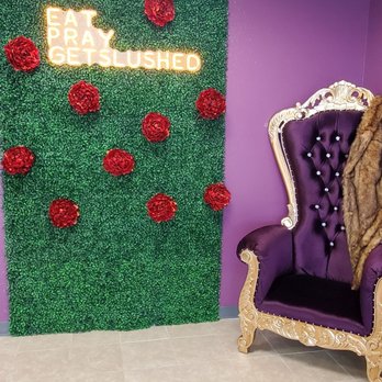 Slushed - One of the many several ops inside. - The image features a decorative wall covered with green artificial foliage, adorned with multiple red roses. A neon sign with the words 'EAT PRAY GETSLUSHED' is mounted on the wall, emitting a bright glow. To the right, there is a purple velvet throne-style chair with an ornate gold frame and button-tufted backrest. The setting appears to be indoors with tiled flooring and a ceiling with visible panels. - Cocktail Bars Near Me - Pflugerville, Texas