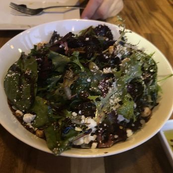 Carmine - A bowl of mixed greens salad containing various leafy greens, cheese crumbles, and nuts is placed on a wooden table. In the background, a person’s hand is visible, along with a plate, fork, and knife, suggesting a dining setting. The image captures a casual meal at a restaurant or home. - Italian Near Me - Newburyport, Massachusetts