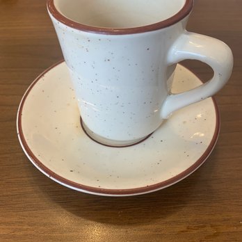 Nancy’s Diner - Coffee! - A white ceramic coffee mug with a brown rim and speckled design is placed on a matching saucer on a wooden table. The mug is empty, and the table surface is smooth with a natural wood grain. In the background, there are utensils and a napkin, indicating a dining setting. - American Near Me - Winnebago, Illinois