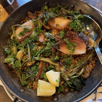 Thistle & Leek - Vegetarian paella - A vegetarian paella served in a black skillet, topped with greens, lemon wedges, and grilled vegetables, on a wooden table. - Gastropubs Near Me - Newton, Massachusetts
