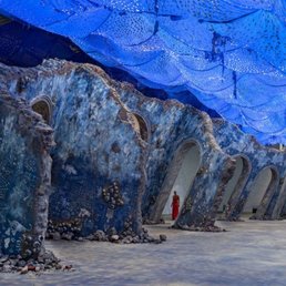 An indoor art installation features a blue fabric canopy overhead and sculptural structures resembling ice or rock formations with arches.