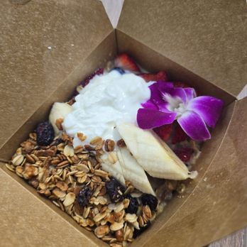 Vanilla Cafe - Crunchy oatmeal - A serving of oatmeal topped with sliced banana, strawberries, a dollop of yogurt, granola, and a purple edible flower is presented in a brown paper container. The dish includes natural ingredients and garnishes, arranged with a variety of textures and colors. - Cafes Near Me - Miami, Florida