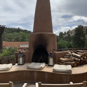 SkyFire - An outdoor fireplace with a tall, adobe-style chimney, surrounded by cushions and lanterns, set against a backdrop of trees and hills. - American Near Me - Santa Fe, New Mexico