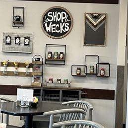 the inside of a store - The image shows the interior of a cafe or coffee shop with a wall decorated with shelves holding jars, small decorative items, and a round sign that says 'SHOP WECKS.' There are also framed pieces of wall art and small vases with flowers on a shelf. In the foreground, there are tables with metal chairs, and a small menu or sign on one of the tables. The decor is modern with a neutral color palette.