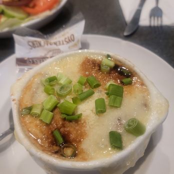 Venus II - Onion Soup - A dish featuring melted cheese, chopped green onions, and a sauce, served in a white ramekin on a plate. The dish appears to be baked, with a slightly browned surface and a creamy texture. The background shows a table setting with a fork, knife, and a partially visible sandwich or burger. The setting suggests a casual dining environment, possibly at a seafood or American restaurant. - Sports Bars Near Me - Marshfield, Massachusetts
