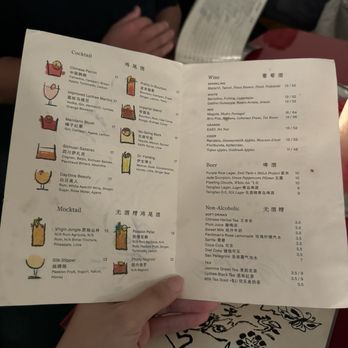 Mala Project - A printed menu from Mala Project in Brooklyn, NY, held by a person at a dining table. The menu includes sections for cocktails, mocktails, wine, beer, and non-alcoholic drinks, with detailed descriptions and prices. The menu features Chinese-inspired drinks and beverages, with illustrations of some drinks. The background shows a dimly lit restaurant setting with other diners and a red table surface. - Chinese Near Me - Brooklyn, New York