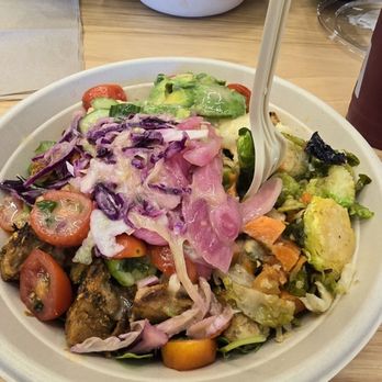 CAVA - Greens & Grains bowl. - A large bowl of mixed salad containing cherry tomatoes, cucumbers, purple cabbage, and grilled protein, with a white plastic fork. - Mediterranean Near Me - Dedham, Massachusetts