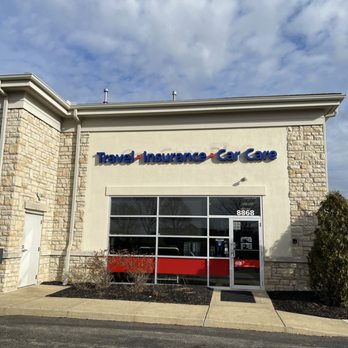 AAA - Powell - A single-story commercial building features a sign displaying 'Travel Insurance Car Care' in blue letters. The address 8868 is visible on the glass door. The facade combines stone and stucco, with a large glass window and door at the entrance. Landscaping includes small bushes and a concrete sidewalk in front. The sky above is partly cloudy, and the overall scene suggests a retail or service business location. - Auto Repair Near Me - Powell, Ohio