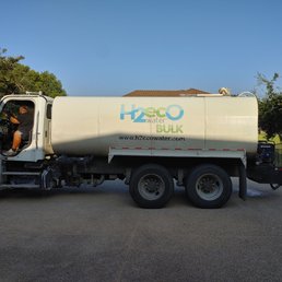 A water delivery truck branded with H2Eco Water and BULK is parked outdoors on a paved surface. The truck has a large cylindrical tank, and the driver is visible sitting in the cab. Trees and a building with a tiled roof are seen in the background, under a clear blue sky.