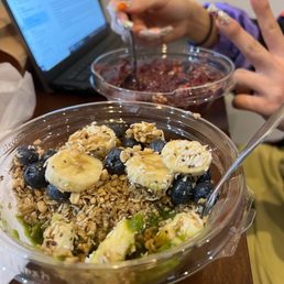 Matcha - Two clear plastic bowls of food are placed on a wooden table. The front bowl contains sliced bananas, blueberries, granola, and a green sauce. The background bowl holds a berry mixture, with a person using a spoon to stir or serve it. A laptop is open nearby, and a person with decorated nails is visible, indicating a casual dining or snack setting.