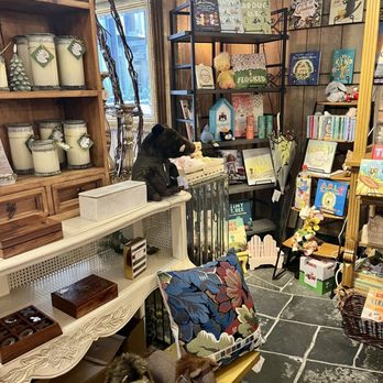Mangel - The image shows the interior of a bookstore or gift shop with wooden and metal shelving filled with books, toys, and decorative items. A black bear plush toy is positioned on a white table, while a small dog rests on a bench nearby. The space has a cozy, eclectic atmosphere with various items like candles, framed pictures, and plush animals. The floor is made of dark stone tiles, and a window provides natural light. The setting appears inviting and well-organized for browsing. - Florists Near Me - Chicago, Illinois