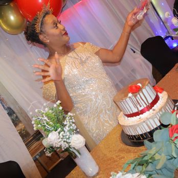 Diana's Bakery - Cute me - A woman dressed in a gold, sequined dress and wearing a crown is celebrating at a party. She is standing near a table decorated with flowers and a two-tiered cake with red and white decorations. The background features balloons in black, gold, and red, along with a sheer curtain and colorful lights, indicating a festive event. - Bakeries Near Me - Phoenix, Arizona