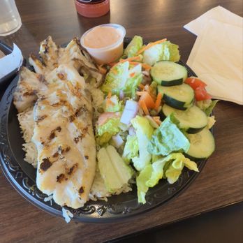 Hooked Seafood Grill - A plate contains grilled fish with visible grill marks, served over white rice. The side salad includes sliced cucumbers, lettuce, shredded carrots, and cherry tomatoes. A small container of pink sauce or dressing is also on the plate. A pink bottled drink is placed behind the plate on a dark wooden table, with a napkin nearby. The setting appears to be a casual restaurant or cafe. - Seafood Near Me - Gilbert, Arizona