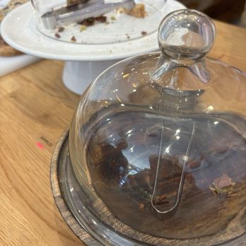 Tinyb Chocolate - samples galore - A glass cake dome with a rounded handle covers a partially eaten chocolate cake with crumbs and small pieces visible inside. The cake is on a wooden serving board, and the setting appears to be a restaurant or cafe with a wooden table surface. In the background, there is a white plate with some food remnants and a glass of water, indicating a dining environment. - Chocolatiers & Shops Near Me - San Mateo, California
