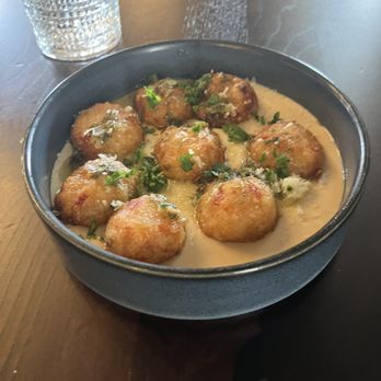 The James - A black bowl contains fried food balls topped with herbs, served in a creamy sauce, with a glass of water nearby. - Wine Bars Near Me - Geneva, Illinois