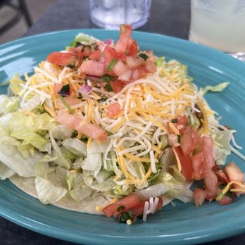Chile Verde - A taco salad featuring shredded cheese, chopped tomatoes, shredded lettuce, and other ingredients is presented on a blue oval plate. The salad is topped with a mixture of shredded cheese and fresh diced tomatoes. In the background, there are two glasses, one with a clear beverage and ice, indicating an outdoor dining environment. The salad appears fresh and colorful, served in a casual restaurant setting. - New Mexican Cuisine Near Me - Columbus, Ohio