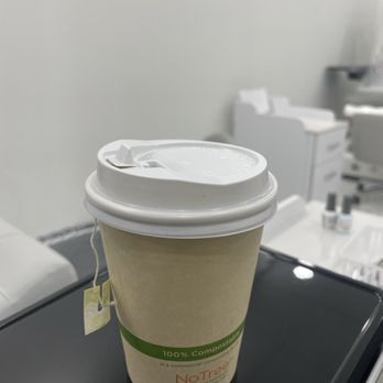 Sofia Nail Spa - Completely hot green tea - A disposable coffee cup made of paper with a white plastic lid is positioned on a black surface. The cup features a green label indicating it is '100% Compostable' and is in a clinical or laboratory environment, with white walls and various medical or laboratory supplies visible in the background. - Nail Salons Near Me - Mount Airy, Maryland