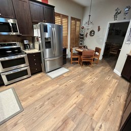 a kitchen with hardwood floors and stainless steel appliances - The image shows a kitchen with dark wood cabinetry, stainless steel appliances such as a refrigerator, microwave, and oven, and granite countertops. There is a small dining area with a round table and four chairs, situated near windows with wooden shutters. The floor is wood, and the space is decorated with wall hangings and a hanging light fixture.