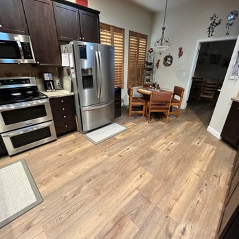 Carpet Exchange - a kitchen with hardwood floors and stainless steel appliances - The image shows a kitchen with dark wood cabinetry, stainless steel appliances such as a refrigerator, microwave, and oven, and granite countertops. There is a small dining area with a round table and four chairs, situated near windows with wooden shutters. The floor is wood, and the space is decorated with wall hangings and a hanging light fixture. - Flooring Near Me - Parker, Colorado