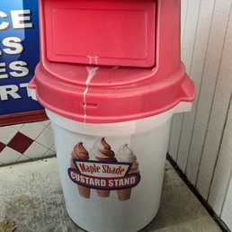 A cylindrical white trash can with a pink lid is positioned outside a business. The lid has a rectangular slot for trash and a drip of liquid is visible on it. The trash can displays a logo for Maple Shade Custard Stand, showing images of ice cream cones. The background includes tiled and wooden wall surfaces, and the ground is concrete with some small debris.