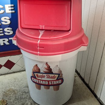 Maple Shade Custard Stand - A cylindrical white trash can with a pink lid is positioned outside a business. The lid has a rectangular slot for trash and a drip of liquid is visible on it. The trash can displays a logo for Maple Shade Custard Stand, showing images of ice cream cones. The background includes tiled and wooden wall surfaces, and the ground is concrete with some small debris. - Ice Cream & Frozen Yogurt Near Me - Maple Shade, New Jersey